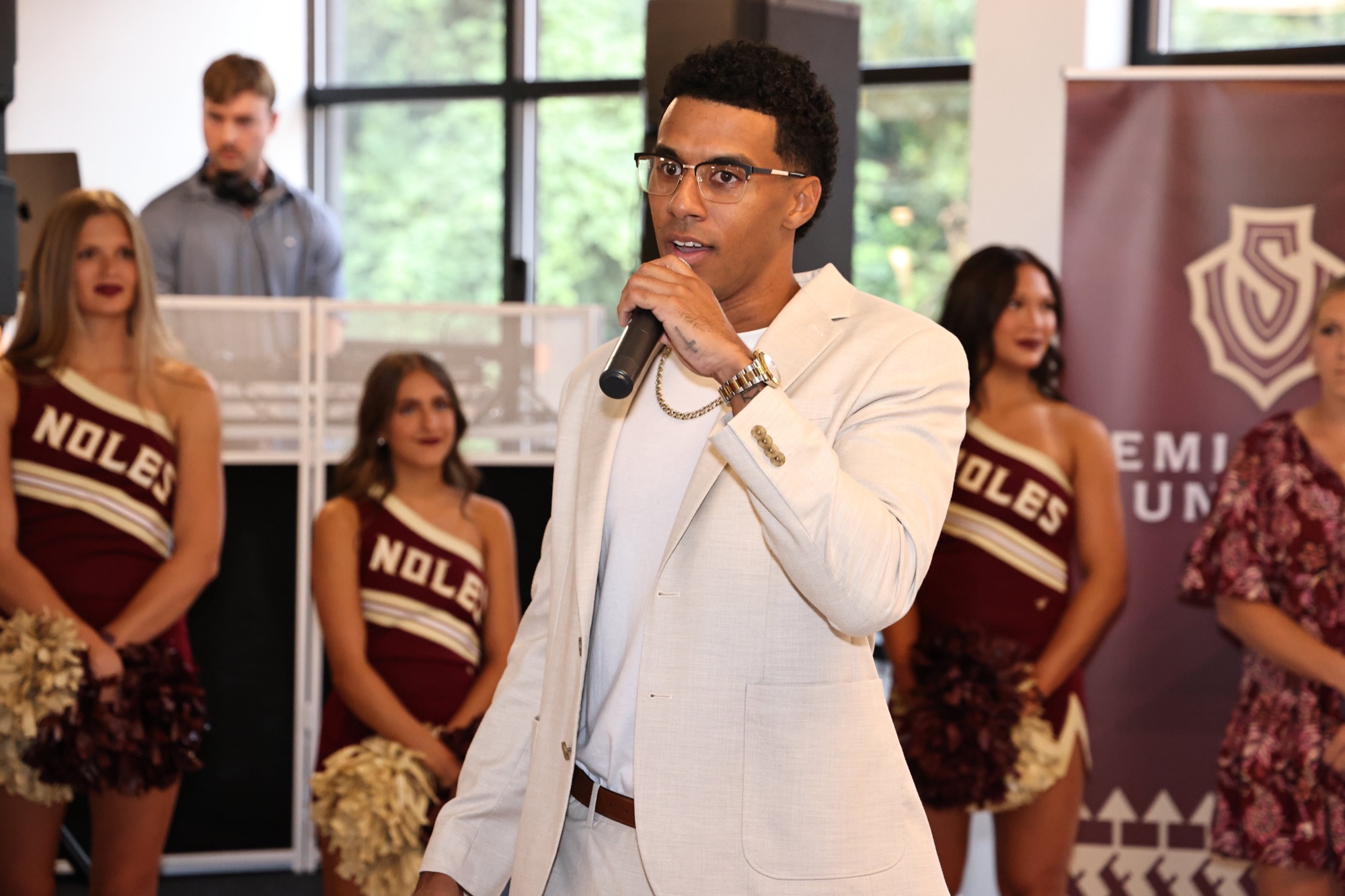 A guest speaking into a microphone at a Seminoles United event, with cheerleaders behind him