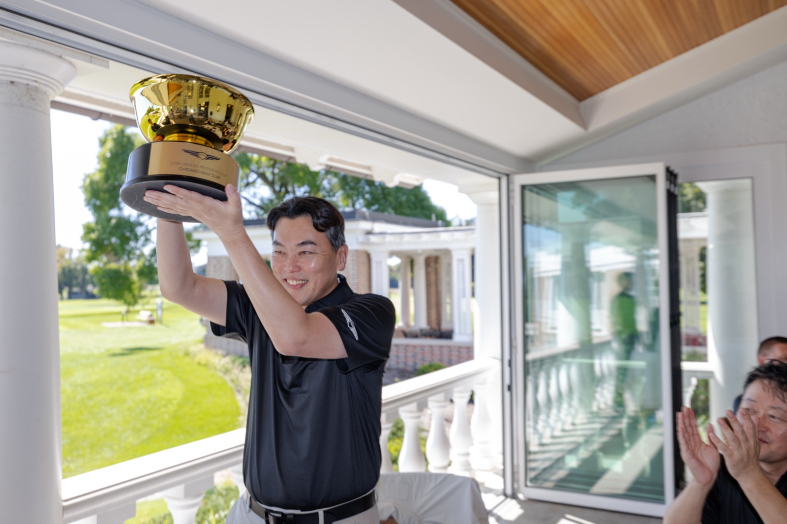 A Genesis guest lifts a gold trophy on the Chicago Golf Club veranda as others applaud