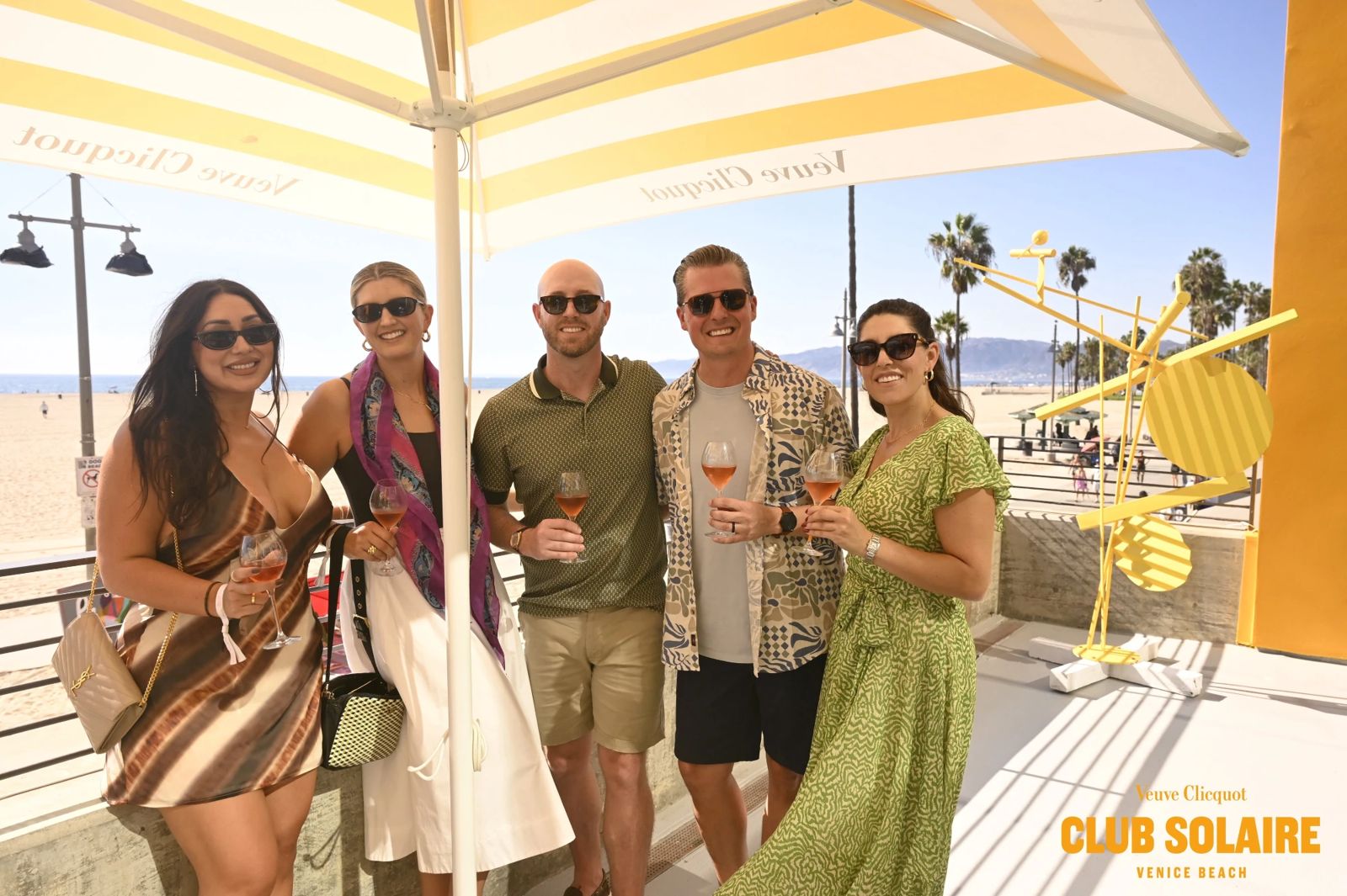 Five Club Solaire guests with glasses of champagne under a striped Veuve Clicquot umbrella above Venice Beach